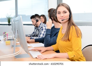 portrait of beautiful and cheerful young woman telephone operator with headset working on desktop computer in row in customer service call support helpline business center with co-worker in background