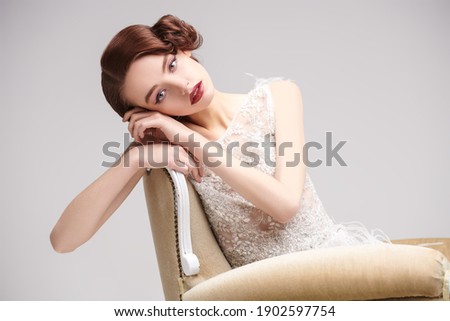 Portrait of a beautiful chic woman posing in a luxury white dress in vintage armchair. Evening makeup and hairstyle of the 20s. 
