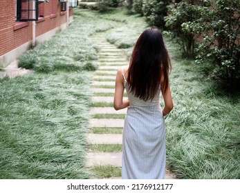 Portrait Of Beautiful Chinese Girl With Black Long Hair In Sundress Walking On Stone Path In Park In Sunny Day. Outdoor Fashion Portrait Of Glamour Young Chinese Cheerful Stylish Woman, Rear View.