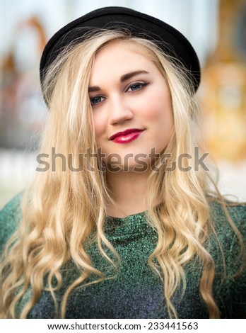 Portrait of a beautiful confident blond blue eyed teenage girl with long hair wearing a bowler hat, glittery t-shirt dark red lipstick standing alone in front of a fairground ride with a white fence.