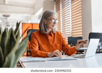 Portrait of beautiful confident senior woman, manager using laptop computer working online sitting in modern office. Business woman wearing stylish eyeglasses checking email. Technology concept  - Powered by Shutterstock