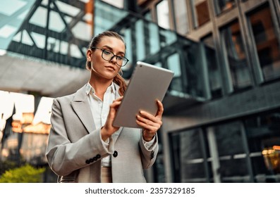 Portrait of beautiful confident successful serious modern adult business woman stylishly dressed in glasses holding digital tablet while standing outside near business center. Technologies, marketing. - Powered by Shutterstock