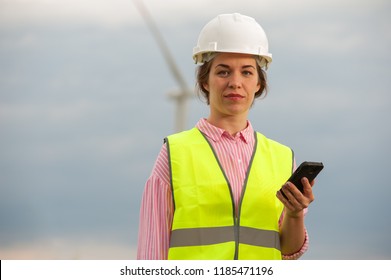 Portrait Of A Beautiful Cute Female Engineer In Shirt In A White Hard Hat Against The Blue Sky And Windmills.