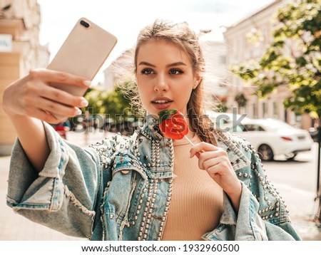 Portrait of beautiful cute model in summer hipster jeans jacket clothes. woman eating lollipop watermelon candy. Woman posing on the street background. Female outdoors taking selfie photos