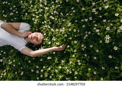 The portrait of beautiful cute woman no makeup in white shirt laying down on green grass with white flowers. Sunny summer day, wild nature, mood vacation, relax. Top view of girl. Be free, love forest