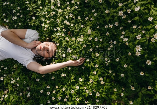 The portrait of beautiful cute woman no makeup in\
white shirt laying down on green grass with white flowers. Sunny\
summer day, wild nature, mood vacation, relax. Top view of girl. Be\
free, love forest