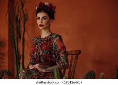 Retrato de una hermosa mujer de cabello oscuro tatuada con peinado coronario trenzado y cabeza floral con elegante vestido de chicharrón sentada en la silla de madera entre los cactus. estilo mexicano. Frida Kahlo 