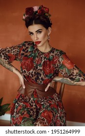 Portrait of beautiful dark-haired tattooed woman with crown braid hairstyle and floral head wreath wearing chic chiffon dress with basque belt  sitting on the wooden chair, hands on her hips.  Mexican