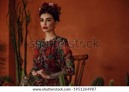 Portrait of beautiful dark-haired tattooed woman with crown braid hairstyle and floral head wreath wearing chic chiffon dress sitting on the wooden chair among cactuses. Mexican style. Frida Kahlo 