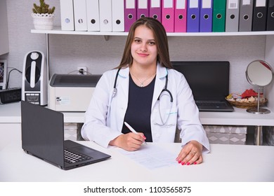 Portrait Of Beautiful Doctor Girl In Office With Laptop. She Sits Right In Front Of The Camera Smiling And Looking Attentive