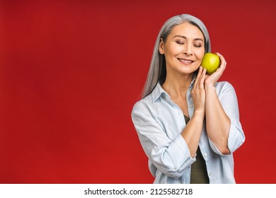 Portrait Of A Beautiful Elderly Mature Senior Asian Aged Woman Holding An Apple, Smiling, Isolated Over Red Background. An Apple A Day Keeps A Doctor Away.