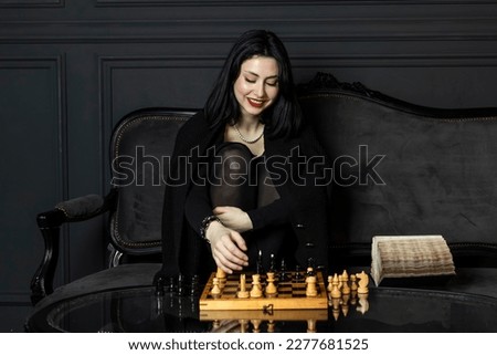 Portrait of a beautiful, elegant girl in black with bright matte red lipstick, sits behind a wooden chessboard, covered herself with a sweater. looks at the chess pieces with a playful expression.