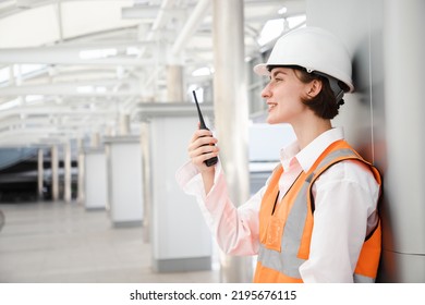 Portrait of beautiful engineering woman using walkie talkie and wear hardhat in front of construction outside work station. Back view of contractor on background of modern buildings.