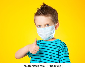 Portrait Of A Beautiful European Boy In A Medical Mask On A Yellow Background. The Child Shows The Sign Super, Class. Big Blue Eye. Epidemics, Pandemics, And The Fight Against Coronovirus.