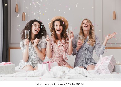 Portrait of beautiful excited women 20s wearing dresses celebrating bridal shower in posh apartment with champagne and falling confetti