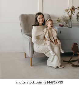 The Portrait Of Beautiful Family Of Mother And Daughter In White Clothes Sitting At Chair At Home. Nice Classic Minimal White Interior. Happy People, Studio Photoshoot, Hugging.