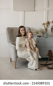 The Portrait Of Beautiful Family Of Mother And Daughter In White Clothes Sitting At Chair At Home. Nice Classic Minimal White Interior. Happy People, Studio Photoshoot, Hugging.