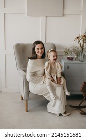 The Portrait Of Beautiful Family Of Mother And Daughter In White Clothes Sitting At Chair At Home. Nice Classic Minimal White Interior. Happy People, Studio Photoshoot, Hugging.