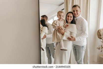 The Portrait Of Beautiful Family Of Mother, Father, Daughter In White Clothes Near Mirror At Home. Nice Modern Minimal Interior. Beige Colours. Happy People, Studio Photoshoot, Hugging.