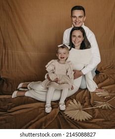 The Portrait Of Beautiful Family Of Three People, Mother, Father And Daughter In White Clothes On Beige Brown Textile Background. Studio Photoshoot, Happy, Hugging, Sitting Down. Winter Sweater.