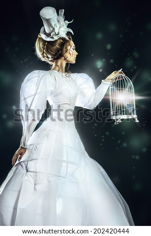 Portrait of a beautiful fashion model in the refined white dress and elegant hat holding birdcage. Black background.