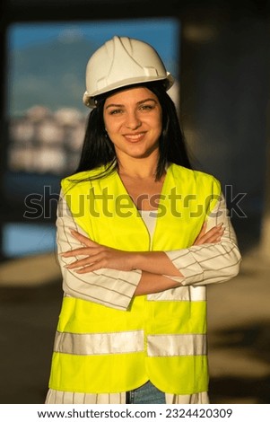 Portrait of a beautiful female designer in a white helmet and yellow vest, looking into the camera. Portrait of an architect standing on a construction site in the rays of the setting sun, copy space.