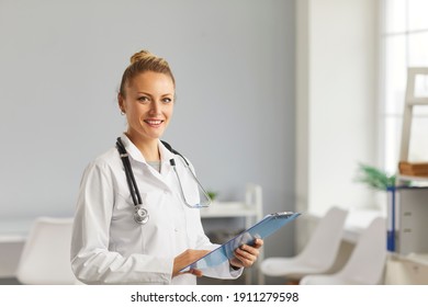 Portrait Of A Beautiful Female Doctor With Documents In Her Hands, Who Is Wearing A White Coat And Standing In A Modern Hospital Room. Healthcare And Medical Concepts.