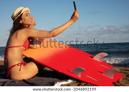 Portrait of beautiful female surfer with her surfboard. Young woman having video call while relaxing at the beach..	
