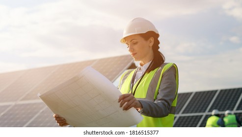 Portrait of beautiful female technologist in uniform with protective helmet. Business woman looking at paper plan placing solar panels.
