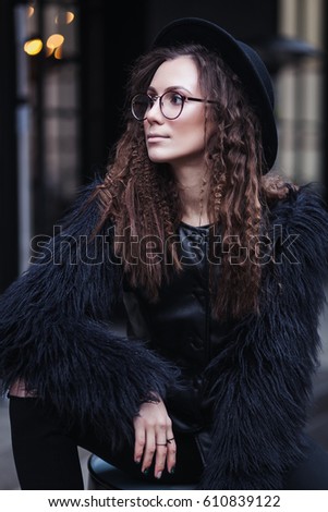 Portrait of a beautiful girl with brown eyes in glasses in a black hat and coat with fur in the cityscape looking away. The girl is like Harry Potter