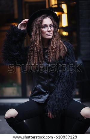 Portrait of a beautiful girl with brown eyes in glasses in a black hat and coat with fur sits on a barrel in the cityscape and looking in camera. The girl is like Harry Potter