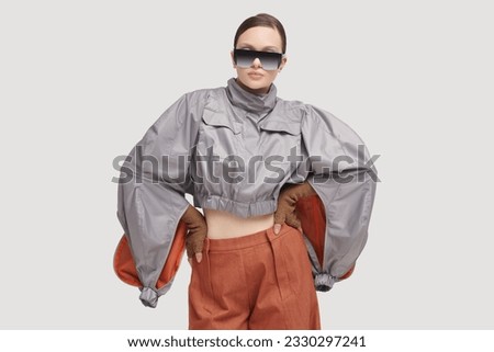 Portrait of a beautiful girl fashion model posing with back in a stylish grey jacket, wide brown pants and mirrored sunglasses on a grey studio background. Futuristic Fashion style. High fashion.