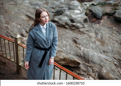 Portrait Of A Beautiful Girl In A Gray Coat On A Gray Background Of A Mountain River. Copy Space