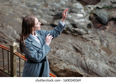 Portrait Of A Beautiful Girl In A Gray Coat On A Gray Background Of A Mountain River. Copy Space