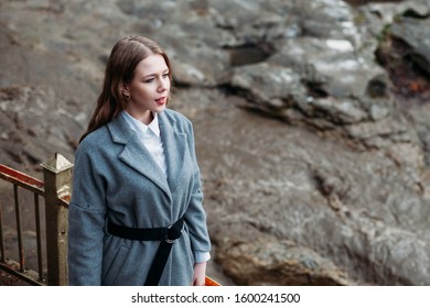 Portrait Of A Beautiful Girl In A Gray Coat On A Gray Background Of A Mountain River. Copy Space