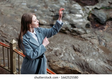 Portrait Of A Beautiful Girl In A Gray Coat On A Gray Background Of A Mountain River. Copy Space