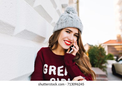 Portrait Beautiful Girl With Long Hair In Marsala Sweater Speaking On Phone On Street. She Wears Knitted Hat And Smiling To Camera