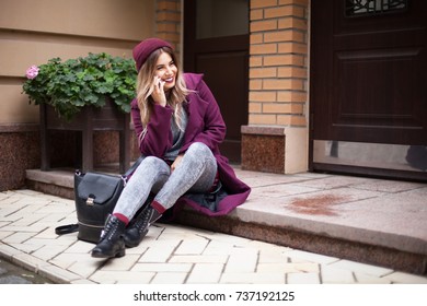 Portrait of a beautiful girl with long hair in a purple coat and gray jeans, talking on the phone in the street near the house. She wears a knitted hat and smiles.