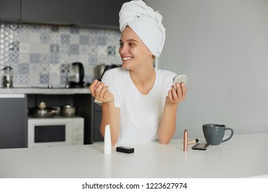 Portrait Of A Beautiful Girl Making Make-up In The Morning, Drinking Tea, Looking And Smiling To Someone A Side, Dressed Casualy And Head Wrapped In A Towel, On A Kitchen Background