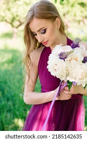 Portrait Of A Beautiful Girl Of Model Appearance In A Purple Fashion Dress And A Bouquet Of Peonies And Lupines. Professional Bridesmaid Makeup. High Quality Photo.