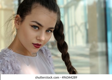 Portrait Of A Beautiful Girl With A Pigtail. She Has Long Brown Hair And Well-groomed Skin. Portrait Against Glass And Metal.