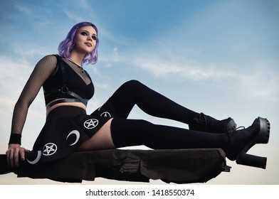 Portrait of beautiful gothic girl against the sky at sunset time. Pastel goth with violet (pink) hair in black clothes