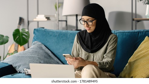 Portrait Of Beautiful Happy Arab Girl In Hijab Working At Home Using Smartphone And Laptop Computer While Sitting On Sofa.