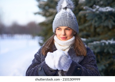 Portrait Of Beautiful Happy Attractive Girl, Young Pretty Woman Is Walking Outdoors In Forest, Snowy Winter Park At Cold Frosty Day, Smiling, Holding Snow, Snowball In Hands In Hat, Scarf And Gloves