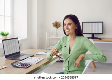 Portrait of beautiful happy female accountant sitting at her workplace in office. Smiling accountant or business analyst in casual clothes sitting in front of laptop , documents and calculator.