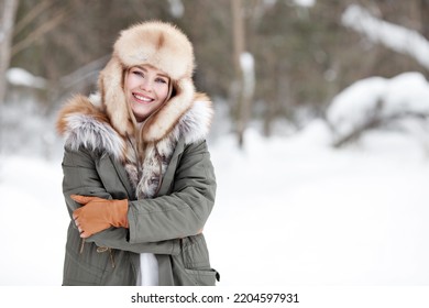 Portrait of beautiful happy girl standing in nature winter snow forest. Smiling woman wearing warm clothes, winter parka jacket, fur hat, leather gloves