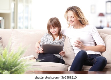 Portrait Of A Beautiful Happy Mother And Her Cute Daughter Sitting In A Beige Sofa Together With A Digital Tablet And Looking Each Other. Modern Mom Teaching Safe Internet Use For Her Preschool Child.