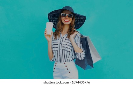 Portrait of beautiful happy smiling woman with shopping bags and coffee cup wearing a black round summer hat on a blue background