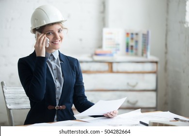 Portrait Of Beautiful Happy Smiling Young Engineer Woman In White Hard Hat Sitting At Office Desk In Architectural Agency. Attractive Model Using Smartphone, Posing With Project Plan. Indoors