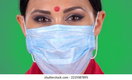 Portrait Of Beautiful Indian Woman In Medical Mask Looking At Camera. Close Up Of A Brunettes Face With Bright Eye Makeup And With Red Bindi Dot On Forehead Posing On Green Screen At Studio.